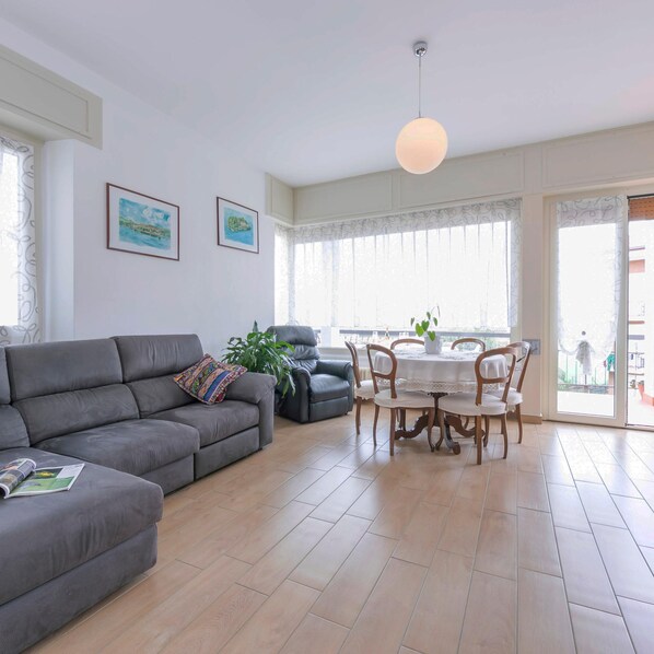 A living room with a grey sofa, a dining table with chairs, and a large window with sheer curtains.