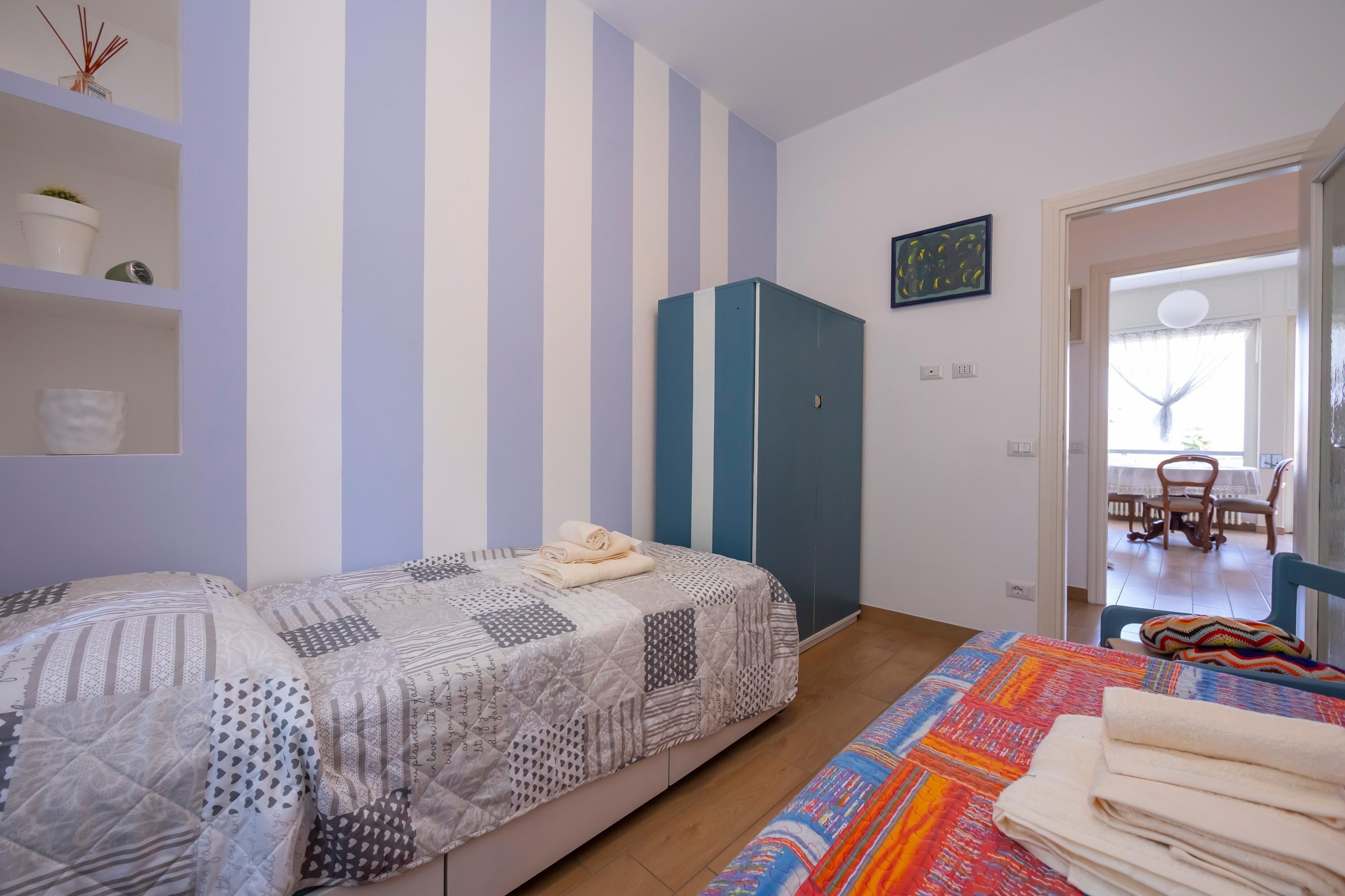 A bedroom with two beds, striped walls, a wardrobe, and a dining area visible through an open door.