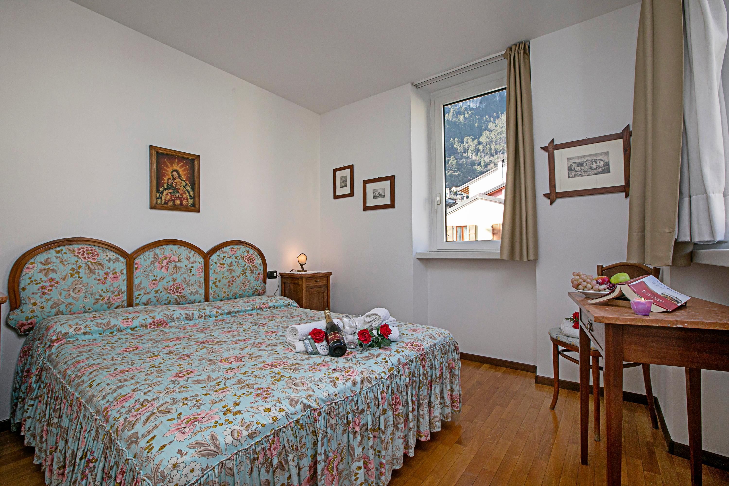 A bedroom with a floral bedspread, a wooden nightstand with a lamp, a window with curtains, and a small table with a book and fruit.