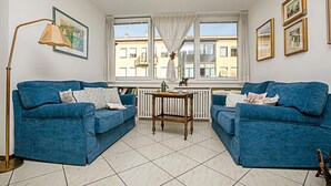 A living room with two blue sofas, a wooden side table, a floor lamp, and a window with curtains. - Riva Centro Apartment (Riva del Garda)