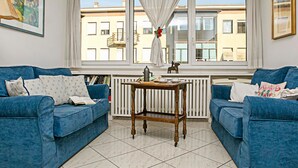 A living room with two blue sofas, a wooden table, and a view of buildings through large windows. - Riva Centro Apartment (Riva del Garda)