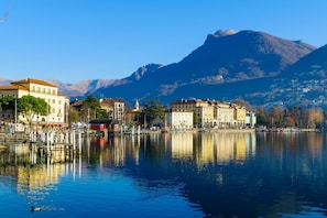 Eine Uferstadt mit Gebäuden entlang des Strandes, ein ruhiger See, der die Bauten spiegelt, und Berge im Hintergrund.