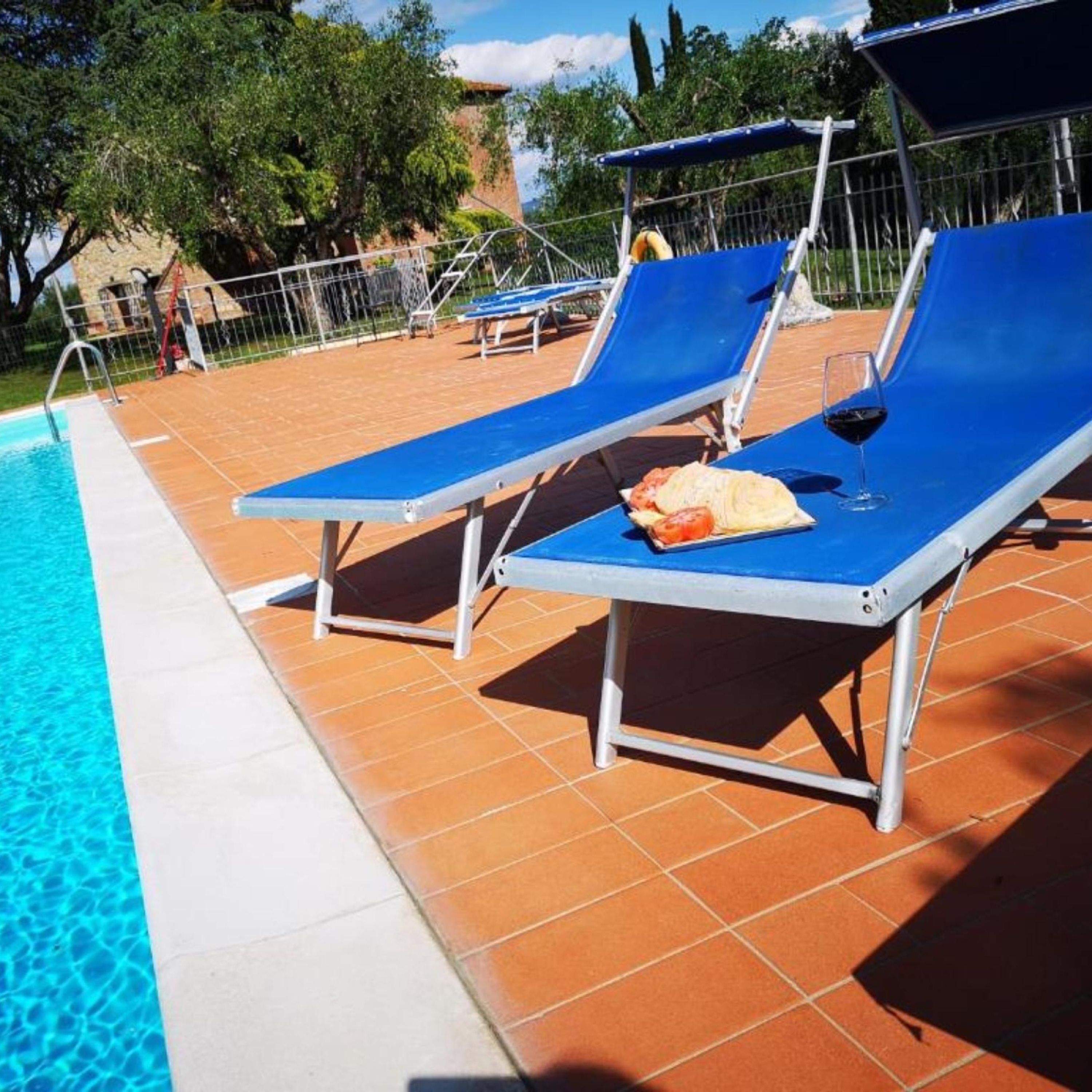 A poolside area with blue lounge chairs, a glass of red wine, and a plate of food.