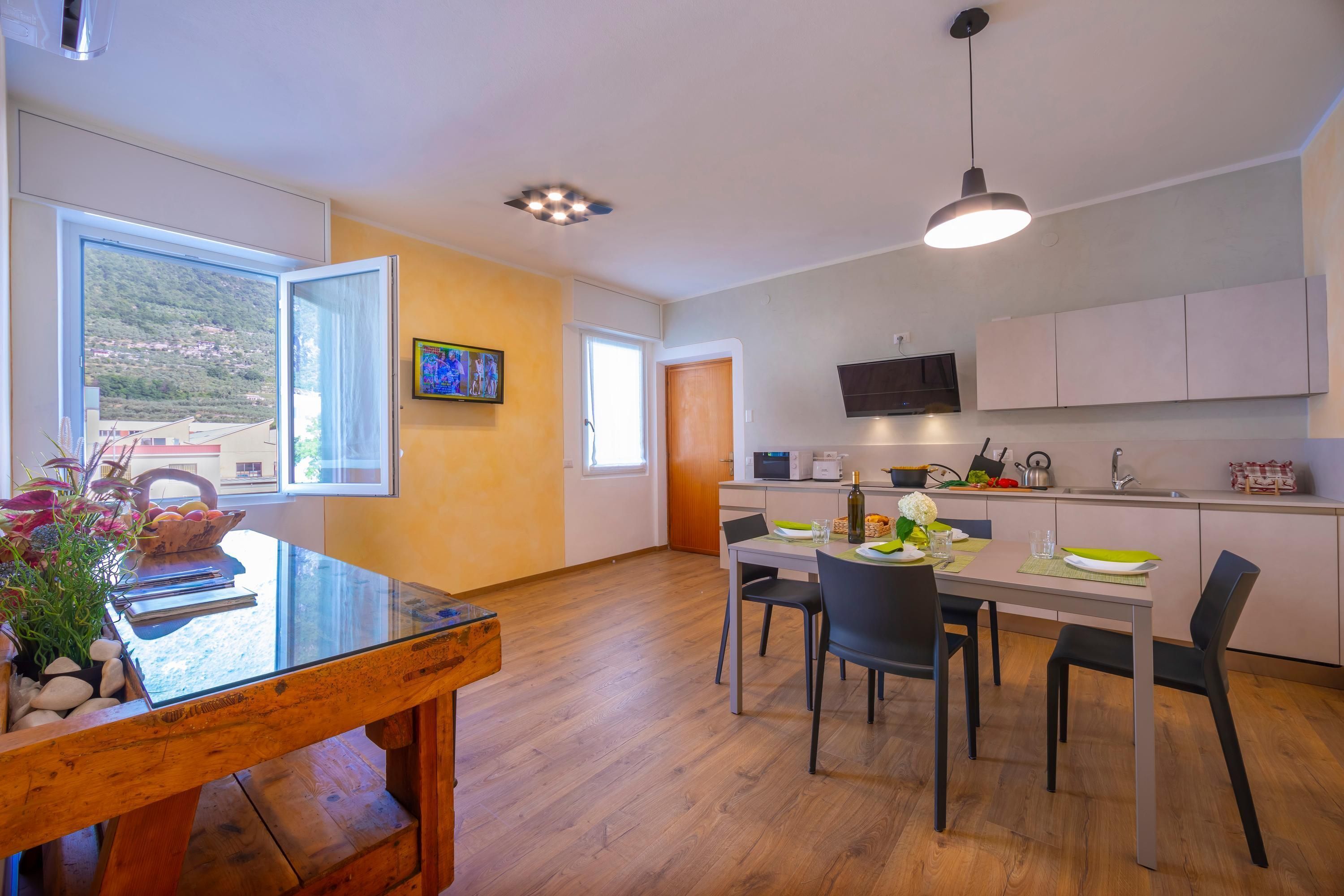 A modern kitchen with a dining area, wooden flooring, and a large window offering a view of a hillside.