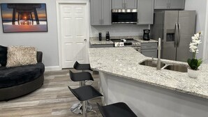 Uma cozinha moderna com armários cinzas, geladeira de aço inoxidável, micro-ondas e bancada de granito. Há um espaço de bar com banquetas e uma orquídea em vaso.
