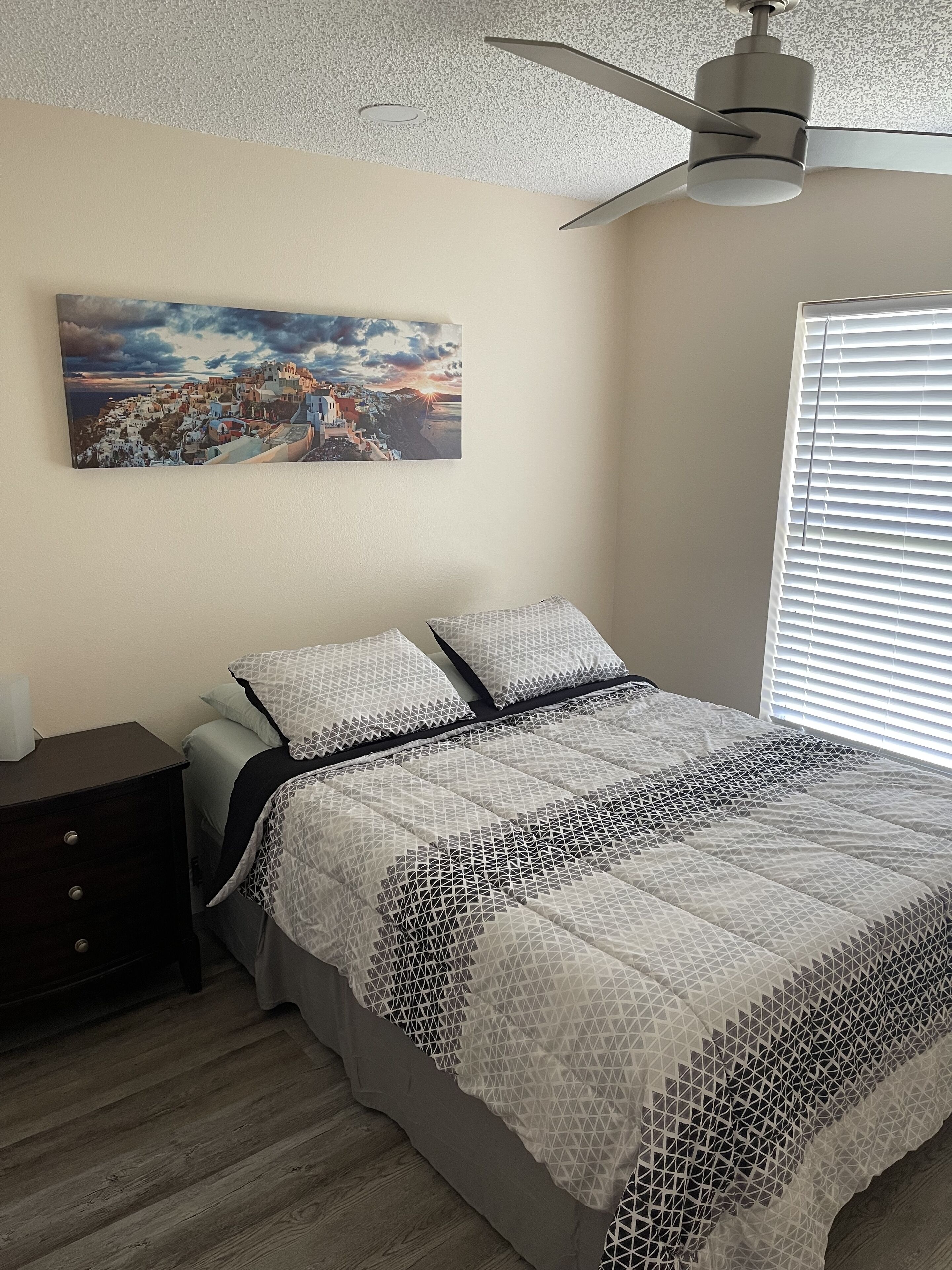 Une chambre à coucher comprenant un lit, une table de chevet, un ventilateur de plafond, une fenêtre avec des stores et un tableau accroché au mur.