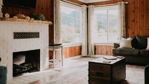 Una sala de estar acogedora con chimenea, paredes de madera, un ventanal con cortinas y un espacio de descanso cómodo.