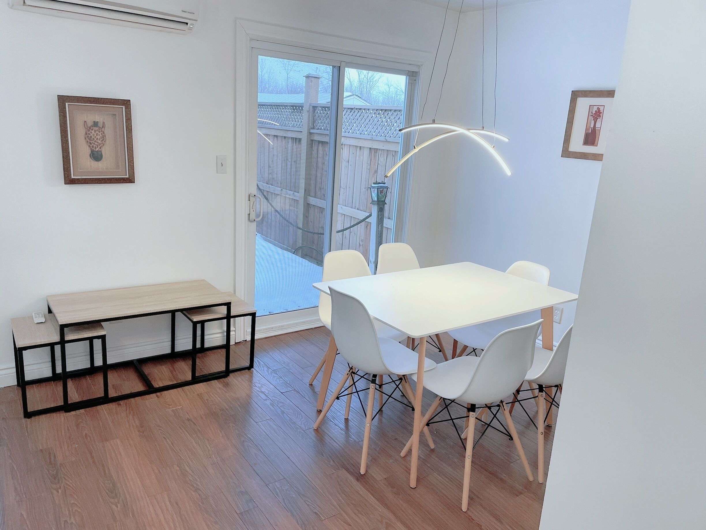 Un espace repas moderne avec une table et des chaises blanches, un banc et une porte-fenêtre coulissante donnant sur une terrasse en bois.