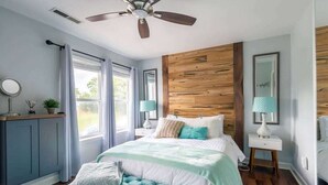 Une chambre à coucher comprenant un lit, un ventilateur de plafond, un mur d’accent en bois, une fenêtre et une table de chevet avec une lampe.