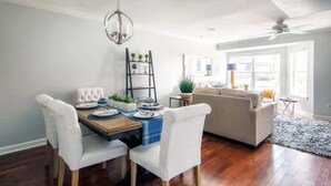 Un coin repas avec une table en bois, des chaises à revêtement blanc, un lustre et une bibliothèque agrémentée de plantes.