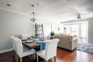Un comedor con una mesa de madera, sillas blancas con tapicería, una araña y una estantería con plantas.