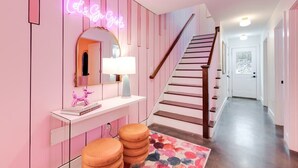 Ein pinkfarbener Flur mit einer Neonreklame, einem Spiegel, einer Treppe und einer weißen Tür am Ende.