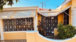 Un edificio de dos pisos con fachada amarilla, un patio adoquinado y un pequeño jardín.