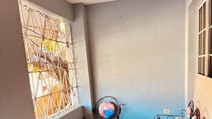 Un pequeño balcón cerrado con piso de baldosas, una mesa metálica, dos sillas metálicas con tapicería floral y una ventana con una reja decorativa de metal.