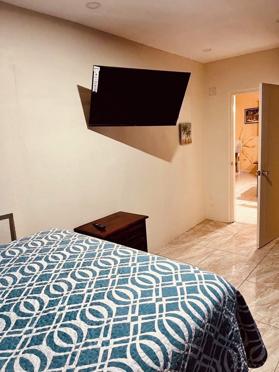 A bedroom with a bed, a TV mounted on the wall, a small bedside table, and a door leading to another room.