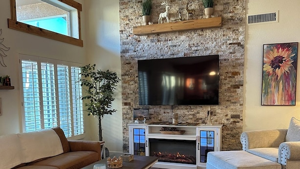 A living room with a stone fireplace, a flat-screen TV, a ceiling fan, and a large window. - Remodeled with access to pools, pickleball, & golf in gated 55+ community! (Goodyear)