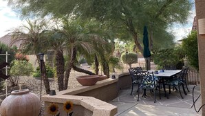 An outdoor patio with a table and chairs, surrounded by plants and a large tree. - Remodeled with access to pools, pickleball, & golf in gated 55+ community! (Goodyear)
