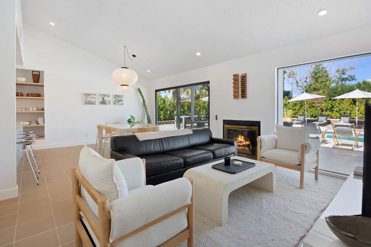 A modern living room with a black leather sofa, white armchairs, a coffee table, and a fireplace. There is a dining area with a table and chairs, and a large window offering a view of the outdoors.