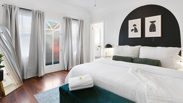 Une chambre moderne avec un grand lit, un banc vert foncé et une vue sur l’extérieur grâce à une porte vitrée.
