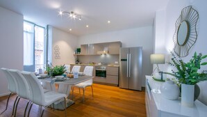 Una cocina moderna con electrodomésticos de acero inoxidable, una zona de comedor con sillas blancas y un ventanal que proporciona luz natural.