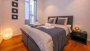 Una cama bien hecha con un edredón gris y blanco, una mesita de noche con una lámpara y una ventana con vistas a edificios.