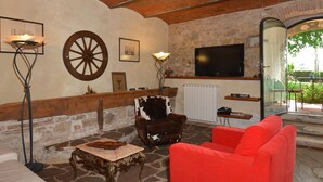 Un salón acogedor con suelo de piedra, un sofá rojo, un sillón de madera y un televisor instalado en una pared de piedra.