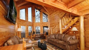 A log cabin interior with a wooden staircase, a leather sofa, a dining table, and a television mounted on the wall. - Smoky Treehouse Lodge #614 (Sevierville)