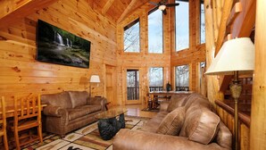 A log cabin interior with a fireplace, wooden furniture, and a large window overlooking a deck. - Smoky Treehouse Lodge #614 (Sevierville)