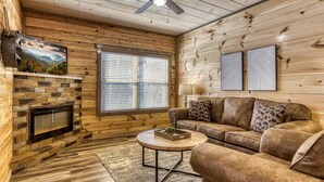 A cozy cabin interior with a stone fireplace, a leather sofa, a ceiling fan, and wooden walls. - Sun Splash #342 (Sevierville)