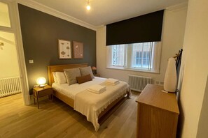 A bedroom with a bed, bedside tables, a radiator, a window with a view, and a nightstand with a lamp. - Central 1 Bedroom Apartment in Farringdon (London)