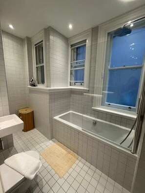 A bathroom with a bathtub, a sink, a window, and a small wooden stool. - Central 1 Bedroom Apartment in Farringdon (London)
