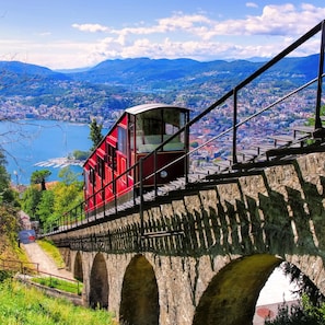 Un funicular rojo sobre un puente de piedra, con un paisaje urbano y montañas al fondo.