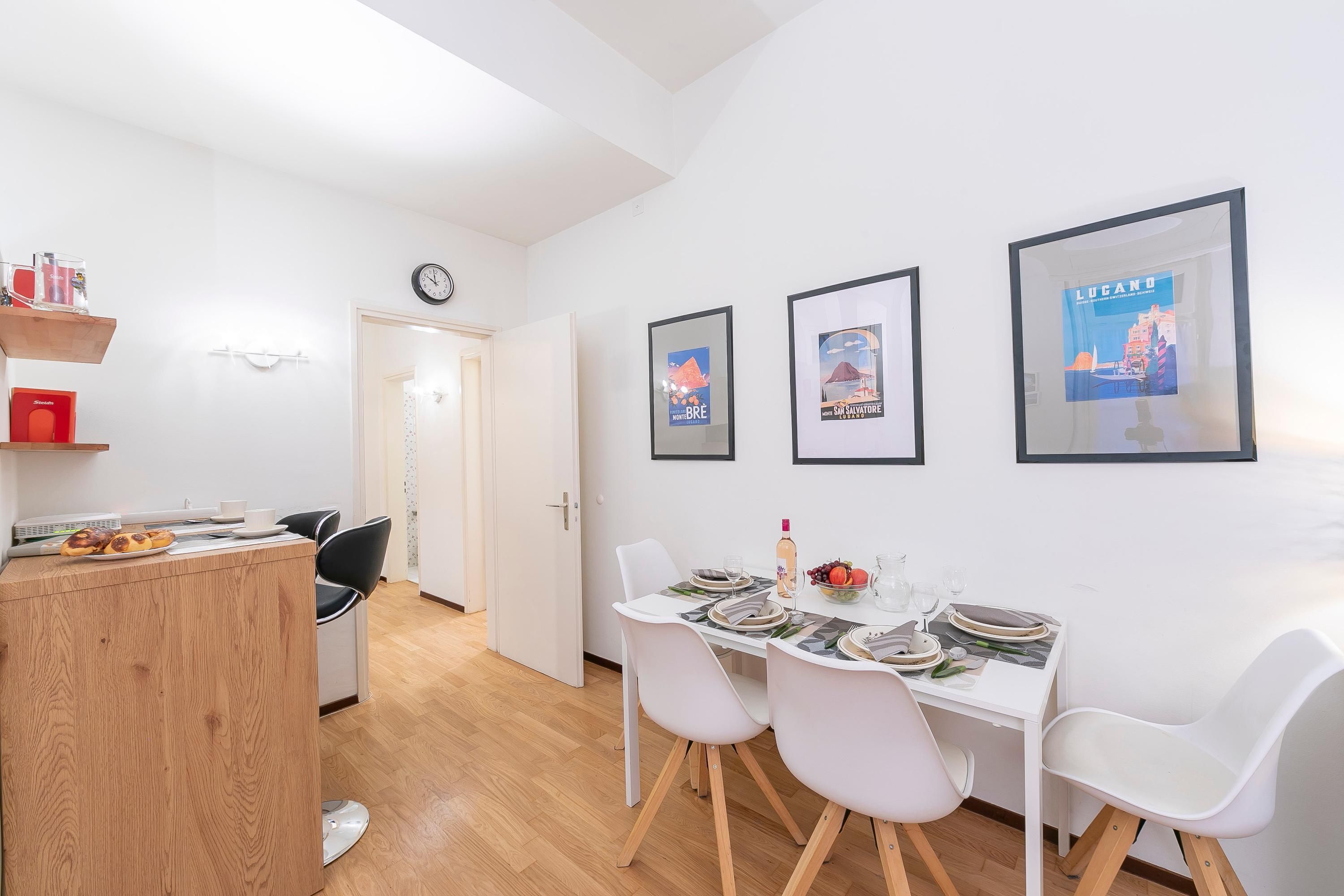 A modern dining area with white chairs and a wooden counter, framed pictures on the wall, and a clock.