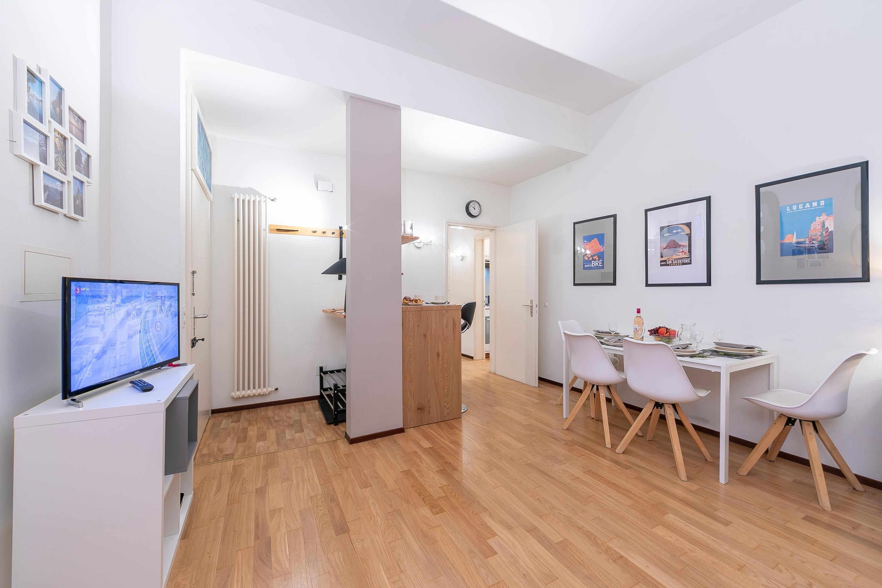 Een moderne, goed verlichte kamer met een houten vloer, een wit bureau met een computer, een televisie en een eethoek met stoelen en een tafel.