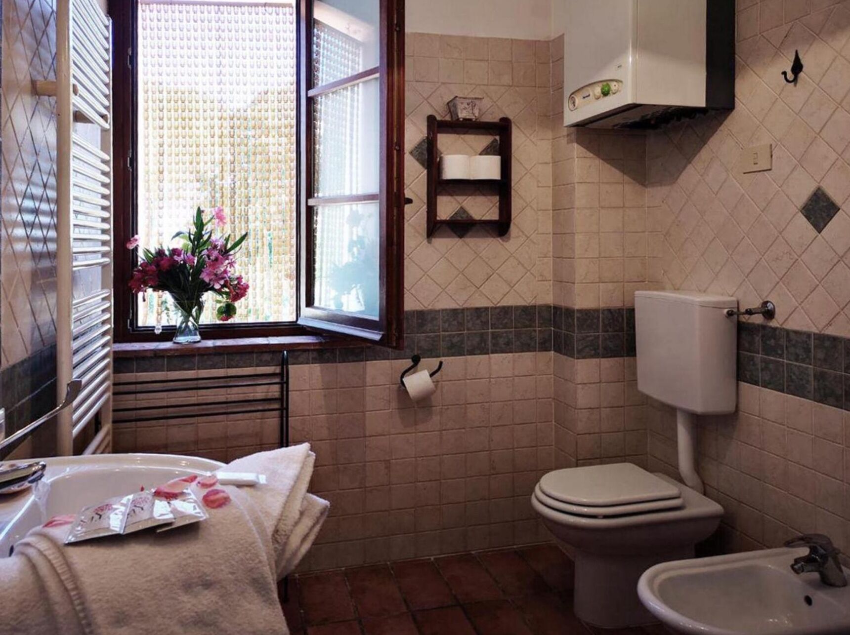 Egy fürdőszoba, amelyben WC, bidé és egy virágokkal díszített ablak található.