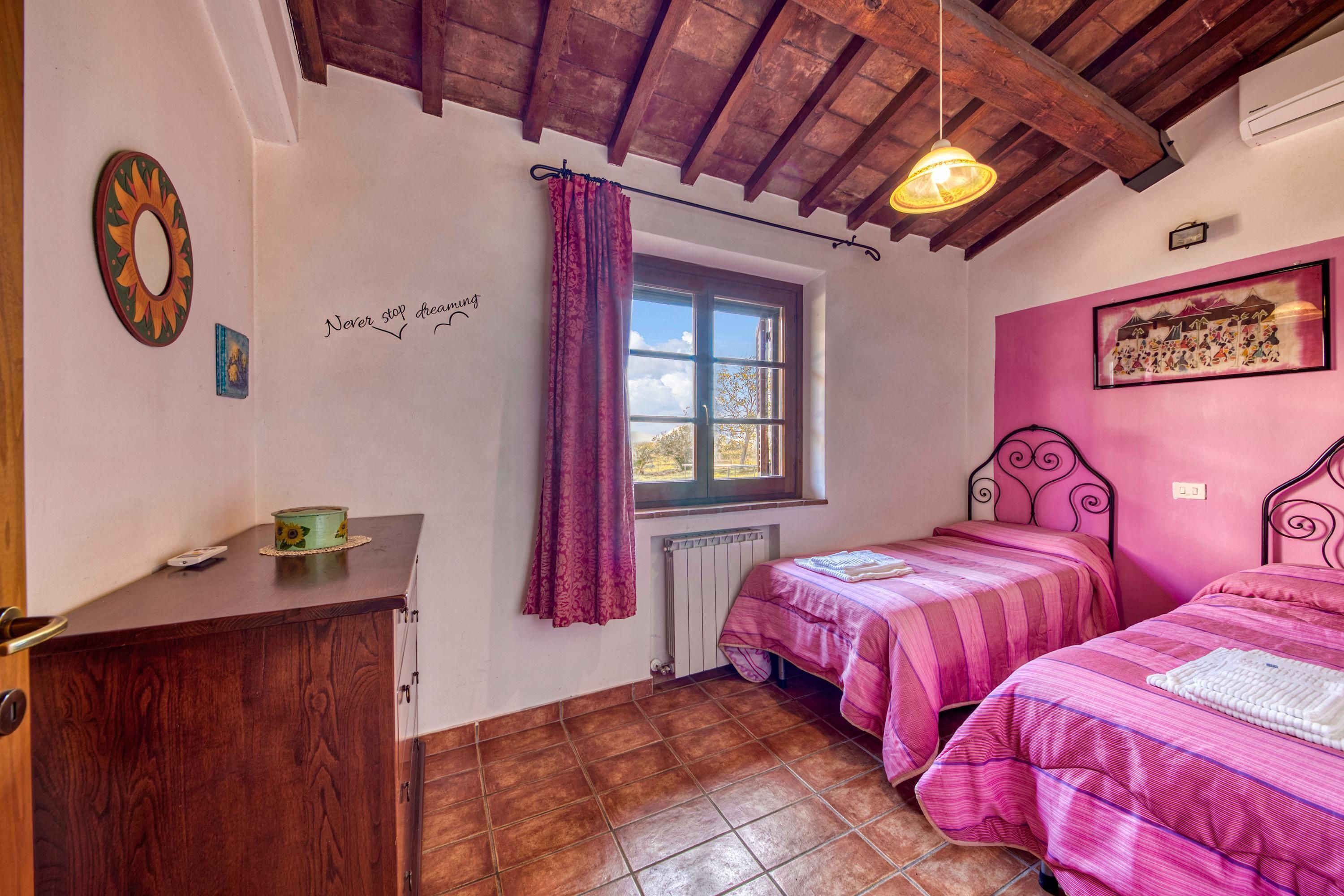 A bedroom with two beds, a wooden ceiling, a window with curtains, a radiator, and a wall with a picture and a quote.