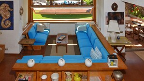 A spacious living area with a large window overlooking a green lawn, a wooden ceiling, and a seating arrangement with blue sofas and a coffee table. - Villa Ada in Ansedonia (Orbetello)