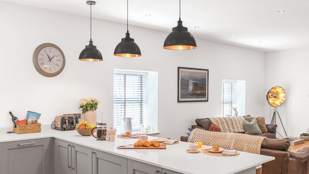 A modern kitchen with a central island, pendant lights, a clock, and a cozy seating area with a couch and floor lamp. - Old Stable (Ulverston)