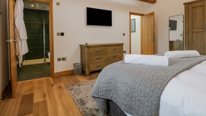 A bedroom with a wooden ceiling, a bed with a grey quilt, a wooden dresser, a television on the wall, and a bathroom with a glass shower. - Old Stable (Ulverston)