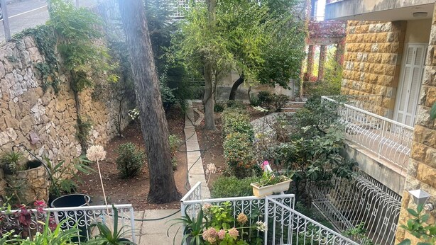 Een stenen muur met een smal gangetje dat uitkomt bij een tuin met potplanten en een metalen hekwerk.