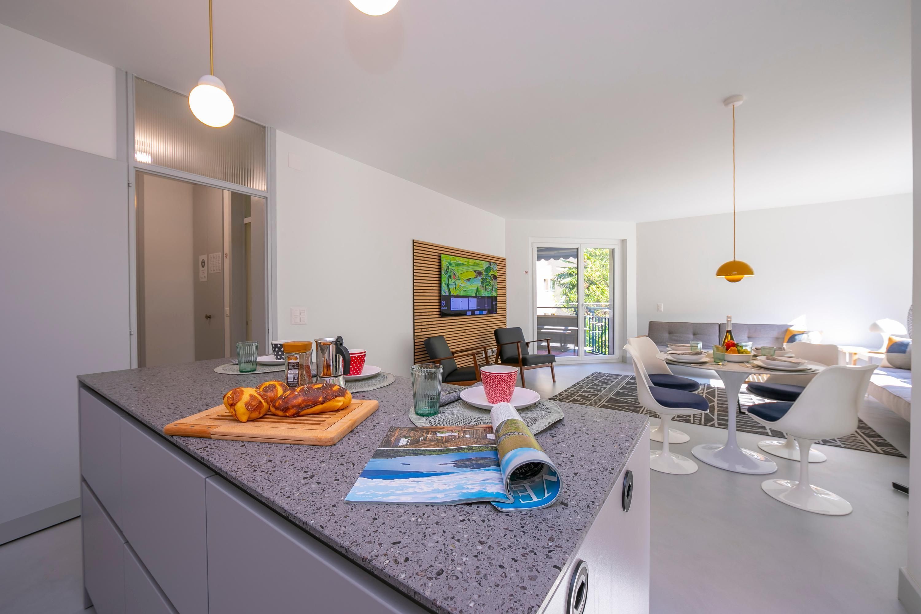 A modern kitchen with a granite countertop, a wooden cutting board with vegetables, and a rolled-up magazine.