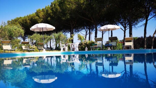 A clear swimming pool with reflections, surrounded by lounge chairs and umbrellas, set against a backdrop of trees. - Juniper apt With Shared Pool Family fun (Castiglione del Lago)