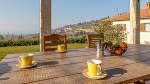 A wooden table with yellow cups and saucers, a coffee pot, and a fruit basket under a covered patio with a view of a hillside. - Panorama Seaview Kolomban (Koper)