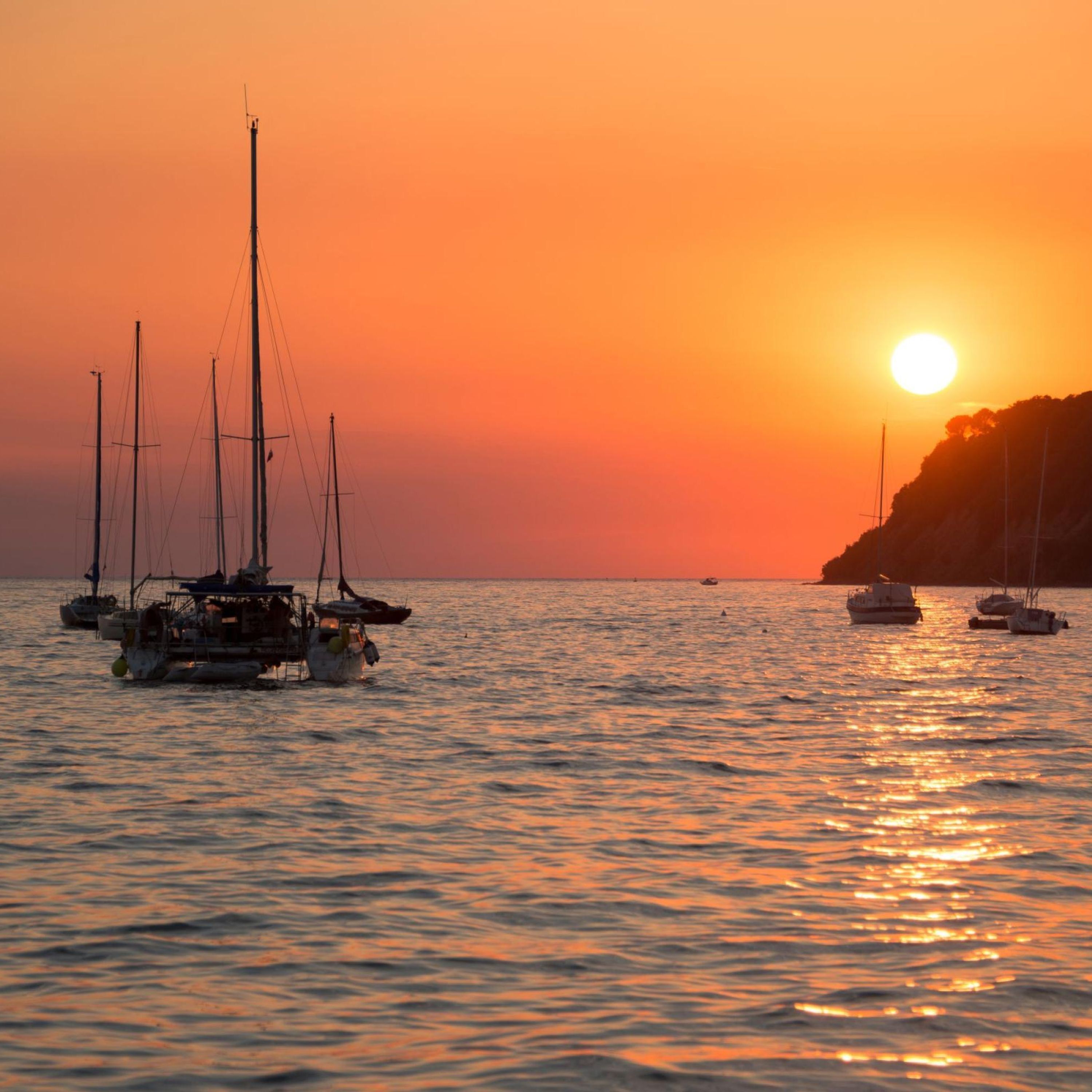 A sunset over a calm sea with several sailboats anchored.