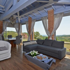 A modern living room with a grey sectional sofa, a white armchair, and a dining table with chairs. A small table with a tray of flowers and snacks is in front of the sofa. The room has a large window with curtains, a chandelier, and a view of greenery outside. - Villa Borgiano in Scansano (Scansano)