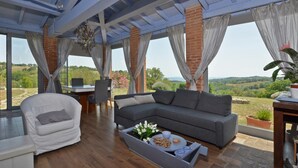 A spacious living area with a grey sectional sofa, a white armchair, and a wooden coffee table with decorative items. Large windows offer a view of the countryside. - Villa Borgiano in Scansano (Scansano)