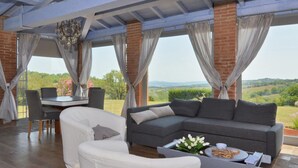 A spacious living area with a large gray sectional sofa, a white armchair, and a coffee table. A dining table with chairs is positioned near a large window offering a view of the countryside. - Villa Borgiano in Scansano (Scansano)