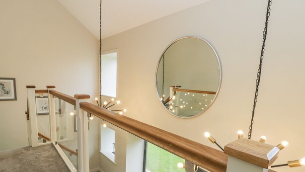 A staircase with wooden handrails and glass railings, leading to a landing with a large round mirror and string lights. - Partridge Place (Hull)