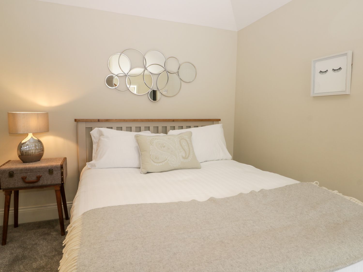 A bedroom with a bed, a nightstand with a lamp, a wall-mounted mirror, and a framed picture.