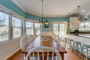 Uma sala de jantar com mesa de madeira e cadeiras brancas, uma cozinha com detalhes em verde-água e grandes janelas.